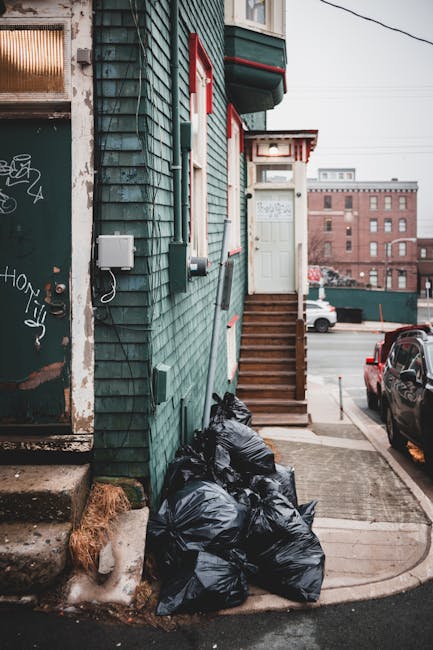 A collection of black garbage bags filled with waste are stacked against the green wooden exterior wall of a building on a residential street. The building features horizontal shiplap cladding painted in a muted green tone, with red and white accents around the small porch area and window frames. A narrow wooden staircase with brown treads leads up to a white door, which is partially visible at the top of the steps. In the background, a parking lot with several cars and a large brick building can be seen, indicating an urban environment. To the left, the edge of a doorway with graffiti and a weathered door frame is visible, alongside utility boxes and cables attached to the building’s exterior. The pavement area is partially covered with a concrete pad, with some patches of snow or ice near the base of the building, suggesting winter or colder weather. The scene captures a snapshot of private waste disposal, possibly an alternative to official rubbish collections, with the focus on the temporarily stored refuse awaiting removal by a waste management service or an independent rubbish clearance company like rubbishclearancebelsizepark.com.