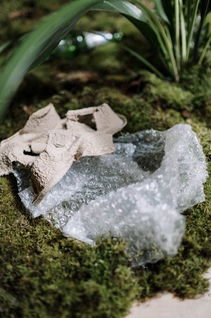 A close-up view of a small patch of mossy ground outdoors, featuring a discarded egg carton made of light brown, textured cardboard and several irregularly shaped pieces of transparent bubble wrap. The egg carton is partially crushed, with some compartments visible, resting on the moss. Surrounding the objects are lush green leaves of plants, with pointed, glossy leaves arching over the scene, suggesting a garden or natural setting. The background is blurred but indicates additional plant foliage, creating a natural environment contrast with the waste materials. The lighting appears soft and natural, highlighting the textures of the moss, cardboard, and bubble wrap, emphasizing the contrast between natural and man-made objects in a setting that implies an informal rubbish accumulation or disposal of packaging waste near garden greenery. This scene may exemplify the importance of responsible waste management and proper disposal methods, supporting themes of private rubbish handling or alternative collection outside municipal services, as seen in some Belsize Park rubbish clearance contexts.