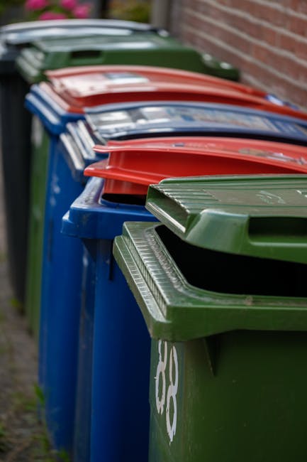 A row of large, rectangular wheelie bins lined up along a sidewalk with a brick wall in the background. The bins are primarily green and blue, with red and dark green lids. The blue bins are positioned in the middle, with the green bins at either end. The lids are closed, and some bins have white symbols or markings on them. The environment appears to be outdoors in a residential area, with the bins placed on a paved surface adjacent to a brick building, possibly for private waste disposal or alternative rubbish collection services. The lighting is natural but slightly subdued, suggesting an overcast day, and the focus is sharp on the bins' front surfaces, gradually blurring towards the back, emphasizing the different colors and textures of the plastic lids and sides.