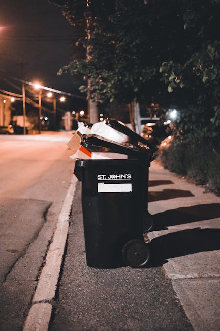 A black wheeled rubbish bin labeled 'St. John's' positioned on a sidewalk near the edge of the street at night. The bin is filled with various rubbish, including cardboard boxes and plastic items, some of which are protruding from the open lid. The scene is illuminated by nearby streetlights, casting warm light and shadows across the pavement and the bin. In the background, there are trees with foliage and a row of parked cars, with additional streetlights visible along the quiet residential street. The environment suggests an urban or suburban area where residents or private waste collection services, such as those offered by rubbishclearancebelsizepark.com, handle waste disposal independently from local authorities. The scene emphasizes the typical setting for private rubbish removal, with the bin positioned ready for collection, reflecting the need for effective waste management outside standard municipality services.