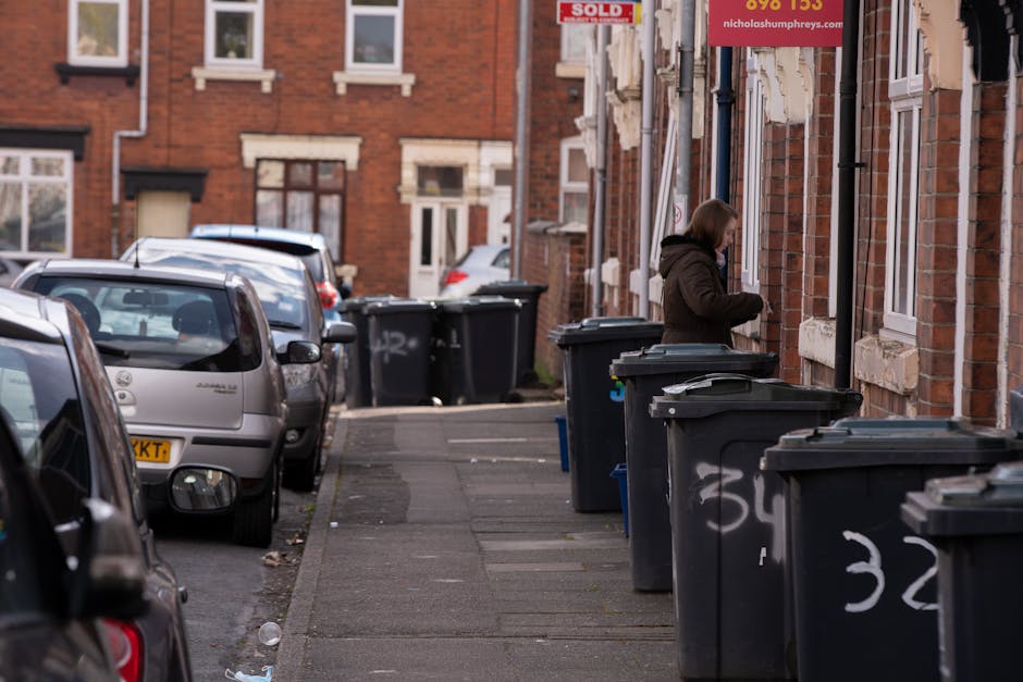 A row of large, rectangular wheelie bins lined up along a sidewalk with a brick wall in the background. The bins are primarily green and blue, with red and dark green lids. The blue bins are positioned in the middle, with the green bins at either end. The lids are closed, and some bins have white symbols or markings on them. The environment appears to be outdoors in a residential area, with the bins placed on a paved surface adjacent to a brick building, possibly for private waste disposal or alternative rubbish collection services. The lighting is natural but slightly subdued, suggesting an overcast day, and the focus is sharp on the bins' front surfaces, gradually blurring towards the back, emphasizing the different colors and textures of the plastic lids and sides.