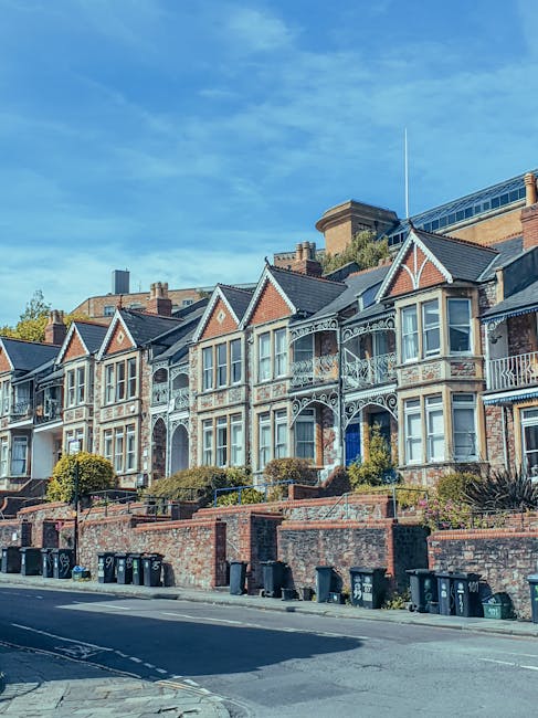 A row of Victorian-style terraced houses featuring ornate ironwork balconies, bay windows, and decorative gables, situated along a paved residential street. The houses have brick facades with light-colored trim and are set behind small, landscaped front gardens bordered by low brick walls. Several black and green wheelie bins are lined up along the curb in front of the properties, indicating waste collection at this location. The sky above is bright blue with some scattered clouds, and the scene is illuminated by natural daylight, highlighting the architectural details of the buildings. The overall setting suggests a typical urban residential area with active rubbish disposal routines, and the presence of bins hints at private waste management or independent waste collection services typical of private residences in the area.